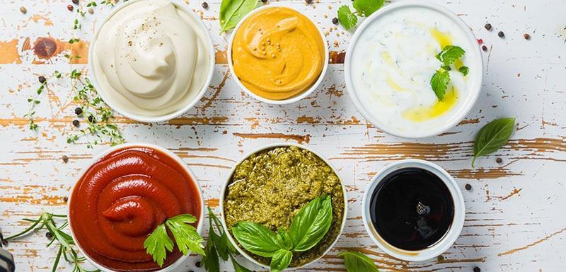 Bowls of sauces and condiments including ketchup, pesto, mustard, soy sauce, mayo, tzatziki